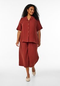 Ensemble deux pièces de couleur rouille comprenant une chemise à manches courtes avec boutons et un pantalon large fabriqué en tissu texturé. Conception simple avec des boutons en laiton.