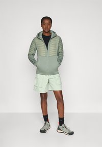 Lys grønn zip-up jakke med en quiltet øvre del, sammen med lys grønn cargo shorts. Modellen har på seg grå tursko og svarte sokker.