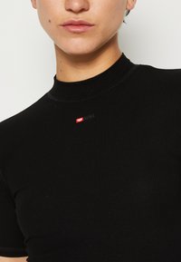 T-shirt en tricot côtelé noir avec un col haut. Présente un petit logo "DIESEL" rouge et blanc sur la partie supérieure de la poitrine. Texture douce.