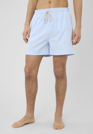 Homme portant un short de bain bleu clair avec un cordon beige, debout pieds nus devant un fond clair uni.