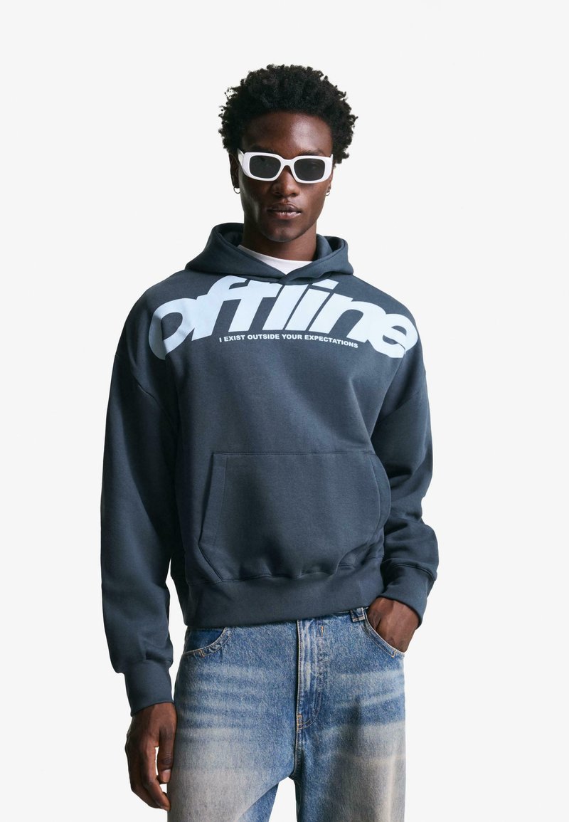 Young man wearing white sunglasses, dark gray hoodie with "offline" text, and light blue jeans, standing with one hand in pocket.