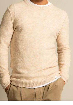 Man in een beige wollen trui over een wit shirt, gecombineerd met beige broek, handen gedeeltelijk in de zakken, tegen een neutrale achtergrond.