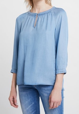 Blusa in denim azzurro chiaro con collo rotondo, apertura a chiave e maniche a tre quarti. Presenta dettagli arricciati sul colletto.