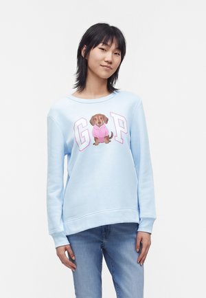 Jeune femme aux cheveux noirs porte un sweat-shirt bleu clair avec un chien de dessin animé en sweat à capuche rose et de grandes lettres G et P, associé à un jean bleu.