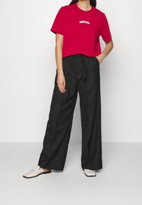 Röd bomull t-shirt med en vit "WOOD WOOD" logotyp, ihopparad med svart byxor med hög midja och strukturerade vita slip-on skor.