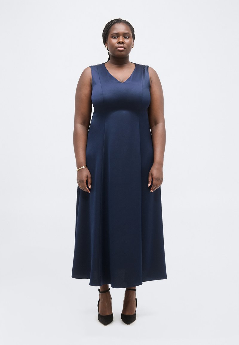 Γυναίκα, όρθια, στραμμένη προς τα εμπρός, φορώντας μια μίντι φόρεμα χωρίς μανίκια σε ναυτικό μπλε και μαύρες γόβες με λουράκι στον αστράγαλο, σε λευκό φόντο.