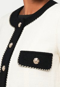 Veste texturée noir et blanche avec un col rond, des boutons dorés et une poche avant avec un élégant détail en tresse.