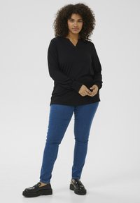 Haut noir à manches longues avec un col en V et des poignets froncés, associé à un jean bleu ajusté et des mocassins noirs à plateforme. Texture lisse et uniforme.