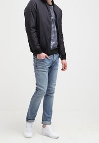 Veste bomber noire avec col côtelé, chemise grise à motifs géométriques, jeans bleu clair et baskets blanches. Tenue simple et décontractée.
