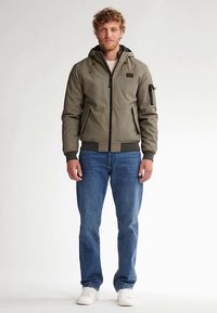 Olivově zelená bunda s kapucí na zip, s bočními kapsami a žebrovanými manžetami, nošená přes krémovou košili a doplněná modrými džínami a bílými botami.