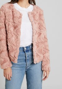 Růžová texturovaná bunda z imitace kožešiny s vlnkovým vzorem, zapínáním na patentky a dlouhými rukávy, nošená přes bílé tričko a modré džíny.