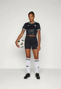 Melna īsā futbolista krekls ar grafisku dizainu, melni šorti un melnas futbola kedas. Baltas zeķes ar trim svītrām, tur rokās futbola bumbu.