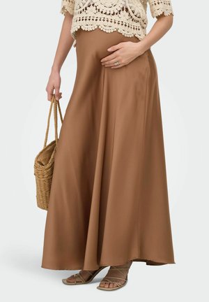 Kvinde i brun satin maxi nederdel, cremefarvet hæklede top, holder en flettet kurvesæk, iført sandaler med stropper, hånd hviler på nederdelen.