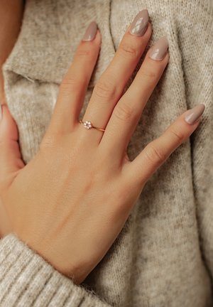 Goldring mit zentralem runden Stein und kleineren Akzentsteinen, getragen an einer Hand gegen einen beigen Pullover. Die Nägel sind in einem hellen Taupe.