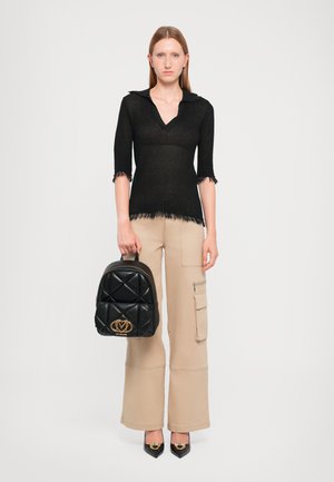 Svart strikket topp med krage og frynsekant, sammen med beige cargo-bukser. Modellen holder en quiltet svart ryggsekk med et gulllogo.