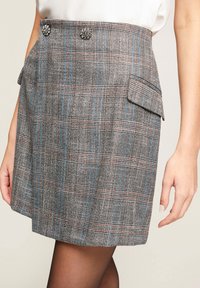 Grijze geruite rok met blauwe en bruine lijnen, twee zijzakken en decoratieve, met juwelen versierde knopen op de taille. Getextureerde stof.