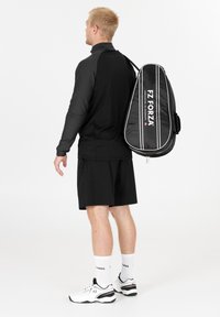 Mustat urheiluvaatteet, joissa on tekstuurilla varustettu pitkähihaine paita, yhdistelevät shortsit ja valkoiset tennarit. Musta mailasäkki roikkuu olkapäällä.