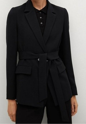 Femme portant un blazer noir sur mesure avec une ceinture, des poches à rabat, et une chemise noire à col en dessous, debout devant un fond uni.