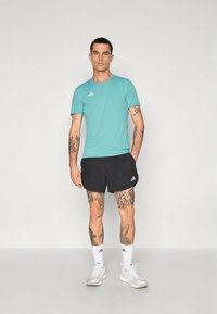 Tyrkysové krátky rukáv športové tričko s logom troch bielych prúžkov, v kombinácii s čiernymi šortkami. Viditeľné tetovania na rukách, biele ponožky a sivé tenisky.