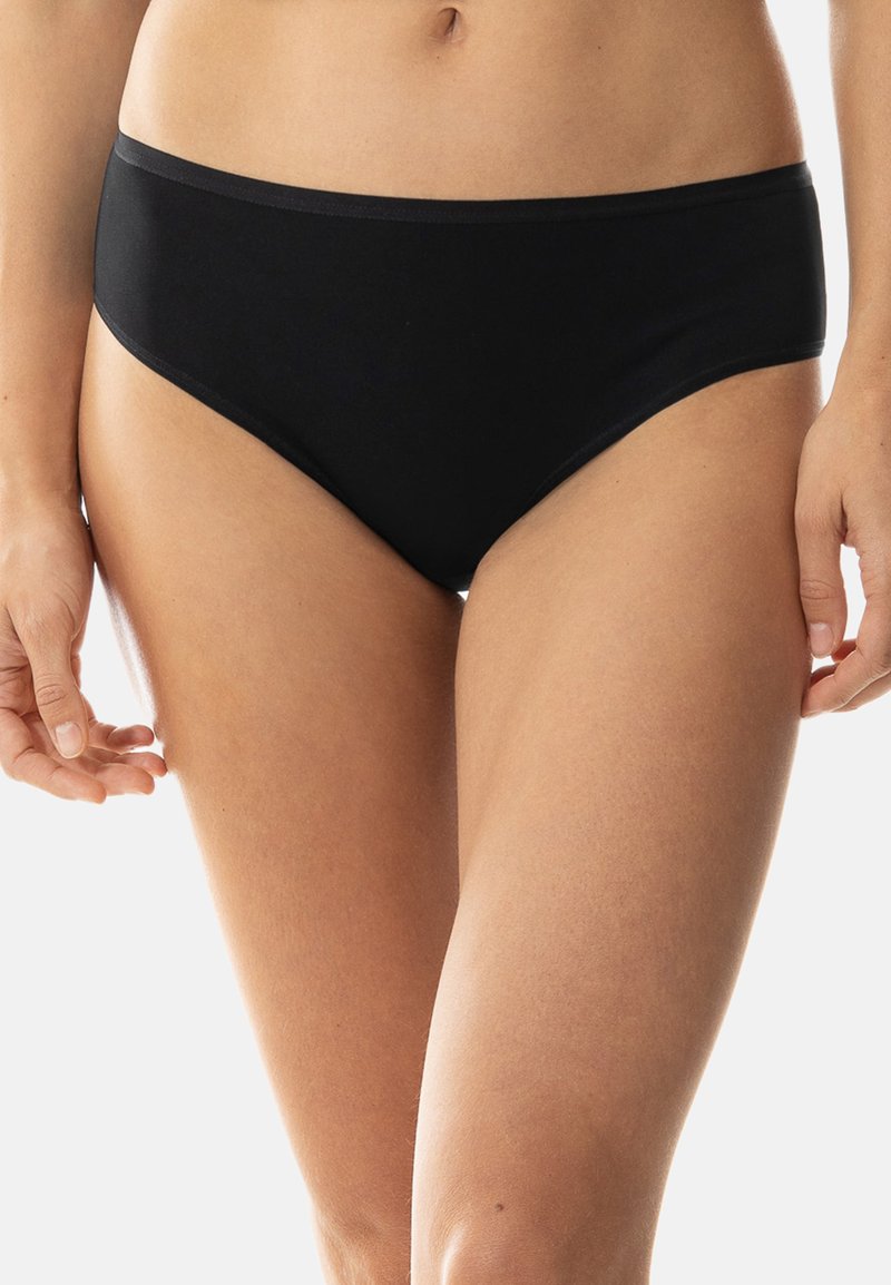 Frau in schlichter schwarzer Bikini-Unterwäsche, die vor einem hellen Hintergrund steht und ihren Mittelbauch und die Oberschenkel zeigt.
