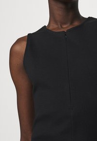 Gros plan sur une personne portant un haut noir à côtes sans manches avec une petite fermeture éclair noire au niveau du cou.