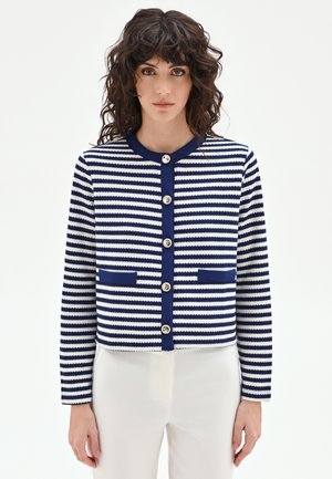 Donna con capelli ricci e scuri che indossa un cardigan abbottonato a righe orizzontali blu navy e bianche e pantaloni bianchi, in piedi davanti a uno sfondo semplice.