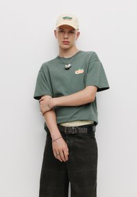 Grünes Baumwoll-T-Shirt mit Pfirsichmotiv, kombiniert mit kurzen Hosen in dunklem Karomuster. Accessoires mit einer beigen Cap und geschichteten Schmuckstücken, einschließlich massiver Ringe.