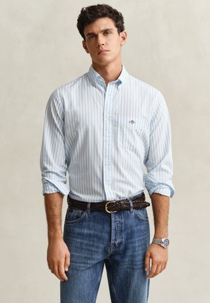 Junger Mann trägt ein hellblau-weiß gestreiftes Hemd mit aufgekrempelten Ärmeln, einen dunkelbraunen geflochtenen Gürtel, blaue Jeans und eine silberne Armbanduhr.