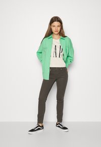 Camisa verde de botões sobre uma t-shirt creme com o logótipo 'GAP', combinada com jeans cinza escuro e sapatilhas pretas com solas brancas.