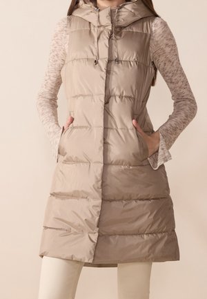 Femme portant un gilet matelassé long beige avec capuche, par-dessus un haut à manches longues marron clair, mains dans les poches du gilet, pantalon de couleur crème visible.