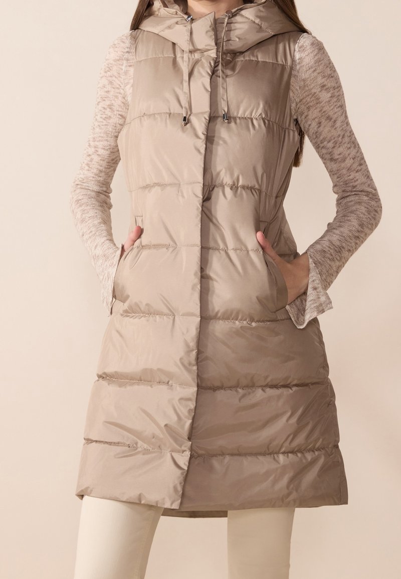Femme portant un gilet matelassé long beige avec capuche, par-dessus un haut à manches longues marron clair, mains dans les poches du gilet, pantalon de couleur crème visible.