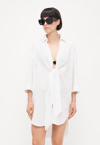 Camicia bianca in lino con nodo frontale, maniche lunghe e vestibilità facile, indossata sopra una maglietta nera. Occhiali da sole scuri e quadrati completano il look.