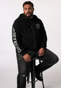 Sudadera negra con cremallera y bolsillos frontales, presenta el texto blanco "COLORADO" en la manga y un emblema en el pecho. Llevada con jeans negros desgastados.
