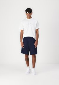 White t-shirt with black text, navy athletic shorts, and white sneakers. Accessories include sunglasses and a visible tattoo on the leg.