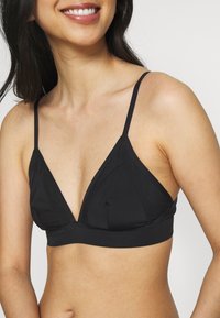 Mulher a usar um sutiã triangular preto com alças finas, a sorrir, mostrada do pescoço até a zona média do torso contra um fundo branco simples.