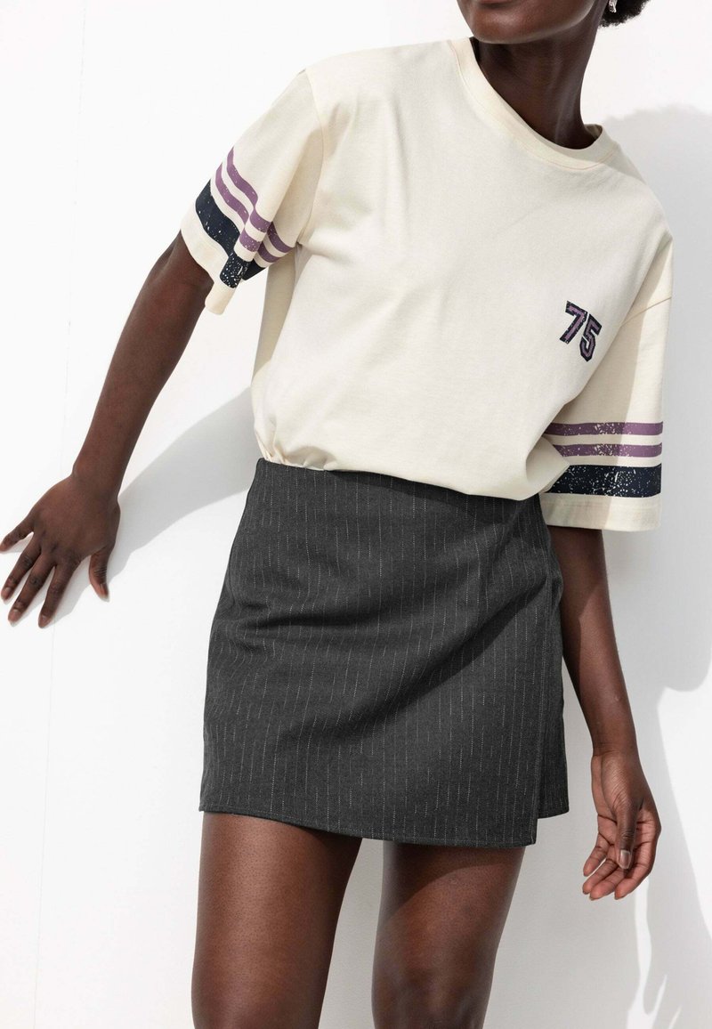 Personne à la peau foncée portant un T-shirt blanc cassé avec des rayures violettes et le numéro 75, associé à une mini-jupe noire à rayures, posant contre un mur blanc.