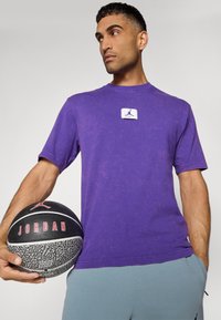 Lilla bomuldstrøje med rund hals, korte ærmer og en hvid broderet logo-patch. Accepteret af en sort og grå basketball.