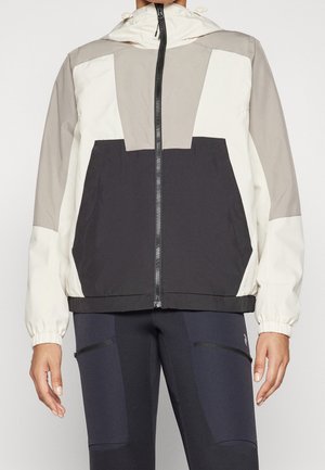 Persona con una chaqueta de bloques de colores en blanco, gris y negro, con cremallera frontal, puños elásticos y leggings negros con cremallera.