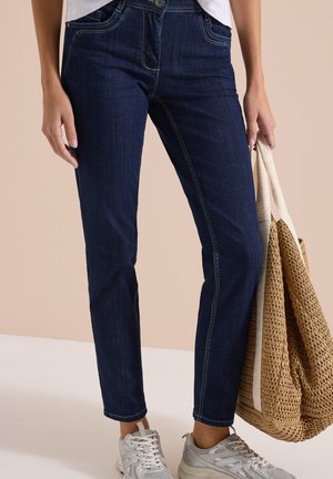 Osoba nosząca ciemnoniebieskie dżinsy i białe trampki, trzymająca dużą beżową plecioną torbę na ramię z kremowymi uchwytami.