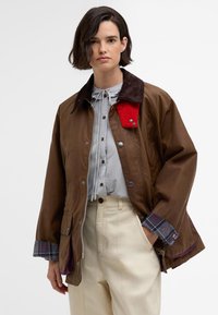 Brown jacket with a corduroy collar, red accent, silver snap buttons, and plaid-lined cuffs. Worn over a light blue shirt and beige pants.