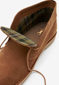 Botas de tobillo de ante marrón con punta redondeada, con un forro de cuadros y suela beige en contraste. Incluye cordones y detalles de costura.