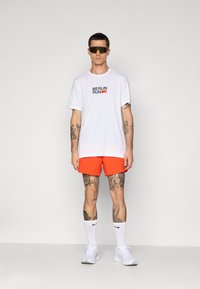 Man wearing white "Berlin Run" t-shirt, bright orange shorts, white Nike socks, white sneakers, dark sunglasses, standing against plain light background.