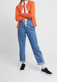 Orange knapp-up cardigan, vit skjorta med röd slips, högmidjade uppvikta blå jeans och svarta sneakers med vita sulor.