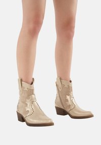 Botines beige con un acabado de ante texturizado, recortes decorativos, acentos con tachuelas y un tacón cuadrado, con una cremallera lateral.