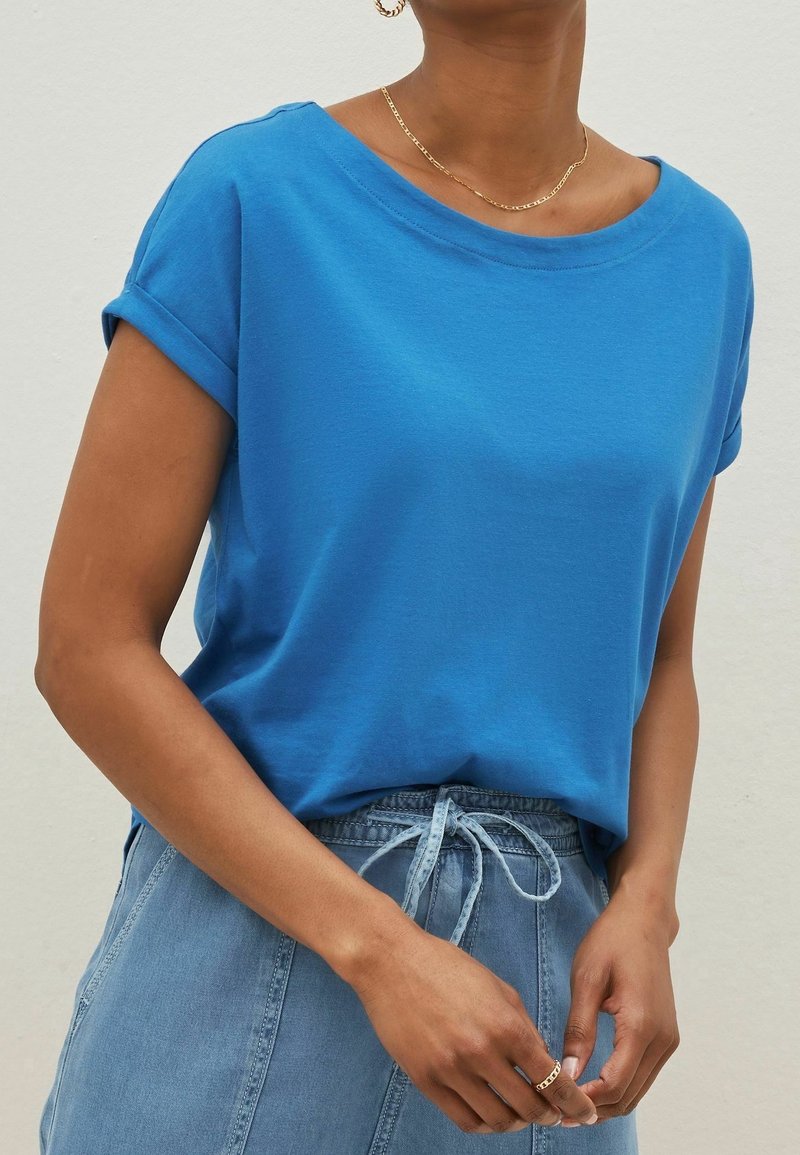 Blå bomulds t-shirt med rullede korte ærmer og bred halsudskæring, kombineret med lyse denim bukser med justerbar talje.
