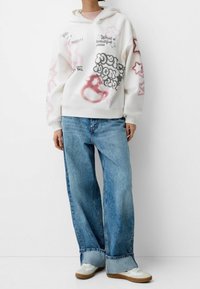 Felpa bianca con diverse grafiche stampate in rosa e nero, abbinata a jeans a gamba larga di un blu chiaro e sneakers bianche con dettagli marroni.