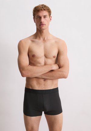 Homme avec les bras croisés, portant un boxer noir avec une ceinture de marque, se tenant devant un fond clair.