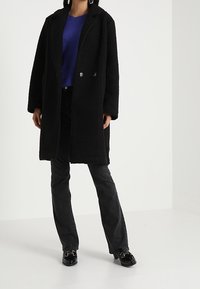 Černý dvouřadý kabát s texturovanou tkaninou, šedé knoflíky, v kombinaci s modrou košilí, černými rozšířenými džínami a lesklými černými botami.
