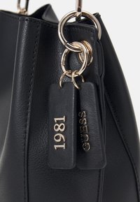 Czarna skórzana torebka z błyszczącymi złotymi elementami, okrągłymi pierścieniami oraz dwoma zawieszkami z wypukłym napisem "GUESS" i "1981".