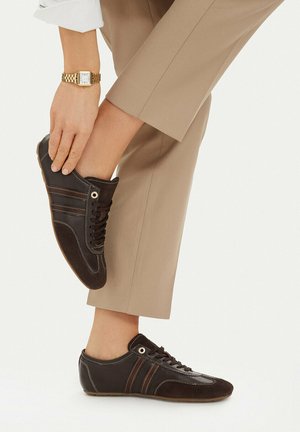 Person iført beige bukser justerer mørkebrune lave sneakers, mens de bærer et gyldent armbåndsur på venstre håndled mod en ensfarvet baggrund.